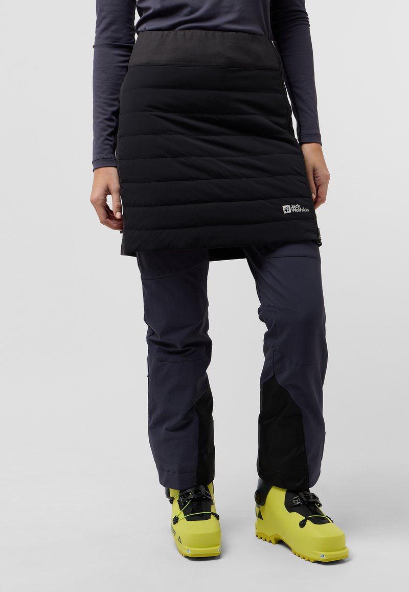 Jupe noire isolée avec un motif matelassé horizontal, associée à un pantalon gris foncé et des bottes de ski jaune vif. Détail du logo sur la jupe.
