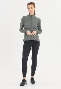 Grå zip-up jakke med ribbet tekstur, tætsiddende design og høj krave, parret med sorte leggings og sorte træningssko.