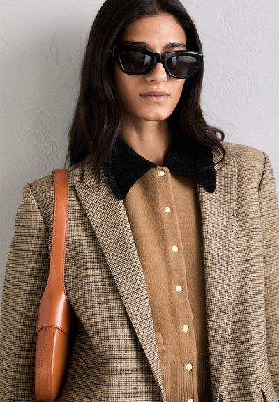 Moteris dėvi per didelius juodus akinius, rudą languotą švarką, smėlinį megztinį su juodu apykakle ir laiko smėlinę odinę rankinę.