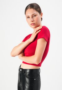 Jongeman met bruin haar in een staart, gekleed in een rode cropped top en zwarte leren broek, poseert met een hand op haar schouder.