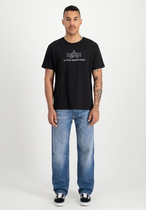 T-shirt en coton noir avec un logo graphique blanc sur la poitrine. Porté avec un jean en denim bleu clair et des baskets noires.