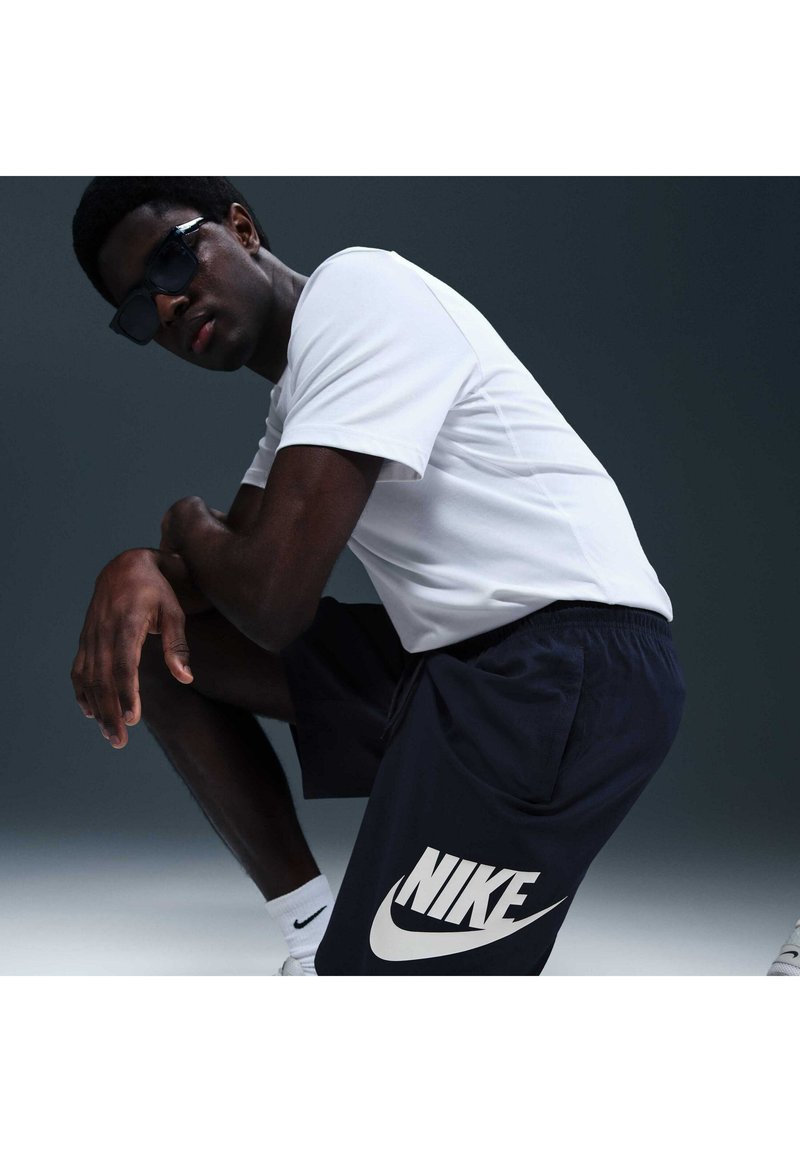 Hombre con gafas de sol negras, camiseta blanca y pantalones cortos Nike negros, agachado contra un fondo gris oscuro.