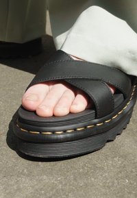 Infradito di pelle nera con cinghie incrociate, suola piatta nera spessa e cuciture gialle a contrasto. Dita dei piedi scoperte visibili sulla superficie superiore.