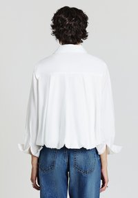 Une personne avec des cheveux courts et bouclés porte une chemise blanche ample à manches longues et un jean bleu, se tenant contre un fond clair uni, le dos tourné vers le spectateur.