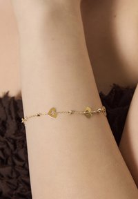 Złota bransoletka z połączonymi małymi serduszkami, z delikatnym łańcuchem i zaokrąglonymi dekoracyjnymi koralikami, spoczywająca na nagim ramieniu.