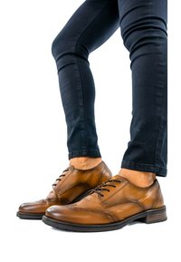 Chaussures de dress en cuir marron portées avec un pantalon sombre ajusté sur un fond blanc, montrant les jambes inférieures et les pieds.