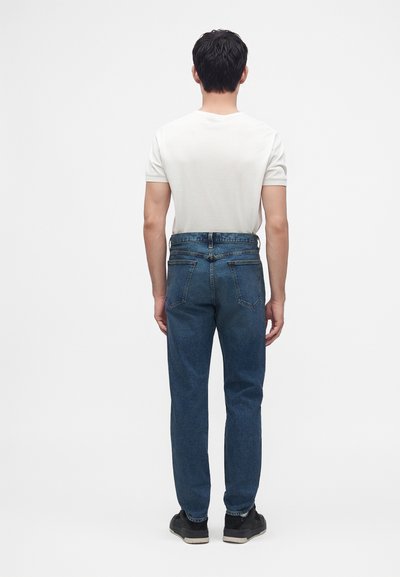 Homme debout, de dos, portant une chemise blanche à manches courtes, un jean bleu et des baskets noires sur un fond blanc uni.