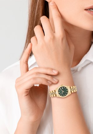 Montre en métal doré avec un cadran vert et un affichage de la date. Présente une lunette texturée et un bracelet à maillons. Portée sur une main à la peau claire.