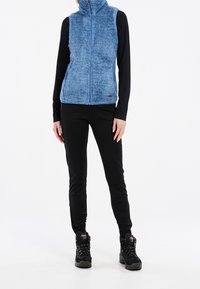 Fleeceweste in Hellblau mit hohem Kragen und Frontreißverschluss. Trägt man über einem langärmligen schwarzen Oberteil, kombiniert mit schwarzen Hosen und schwarzen Stiefeln.