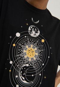 Black shirt with celestial design featuring a sun with a face, moons, planetary orbits, and scattered stars in white and yellow.