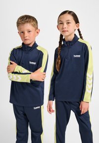 Marinblå och limegröna träningsdräkter med dragkedjekrage. Har reflekterande chevron-accenter och logotyper, tillverkade av slätt tyg.