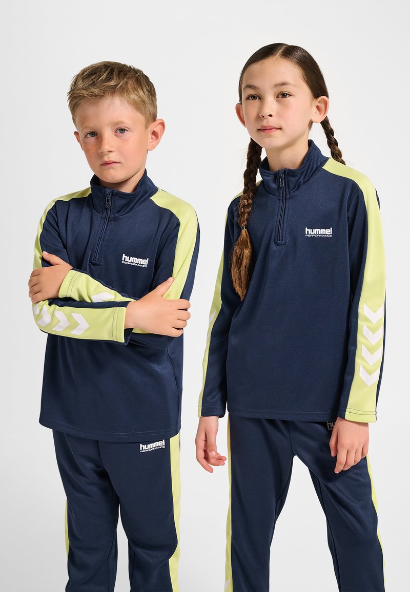 Marinblå och limegröna träningsdräkter med dragkedjekrage. Har reflekterande chevron-accenter och logotyper, tillverkade av slätt tyg.