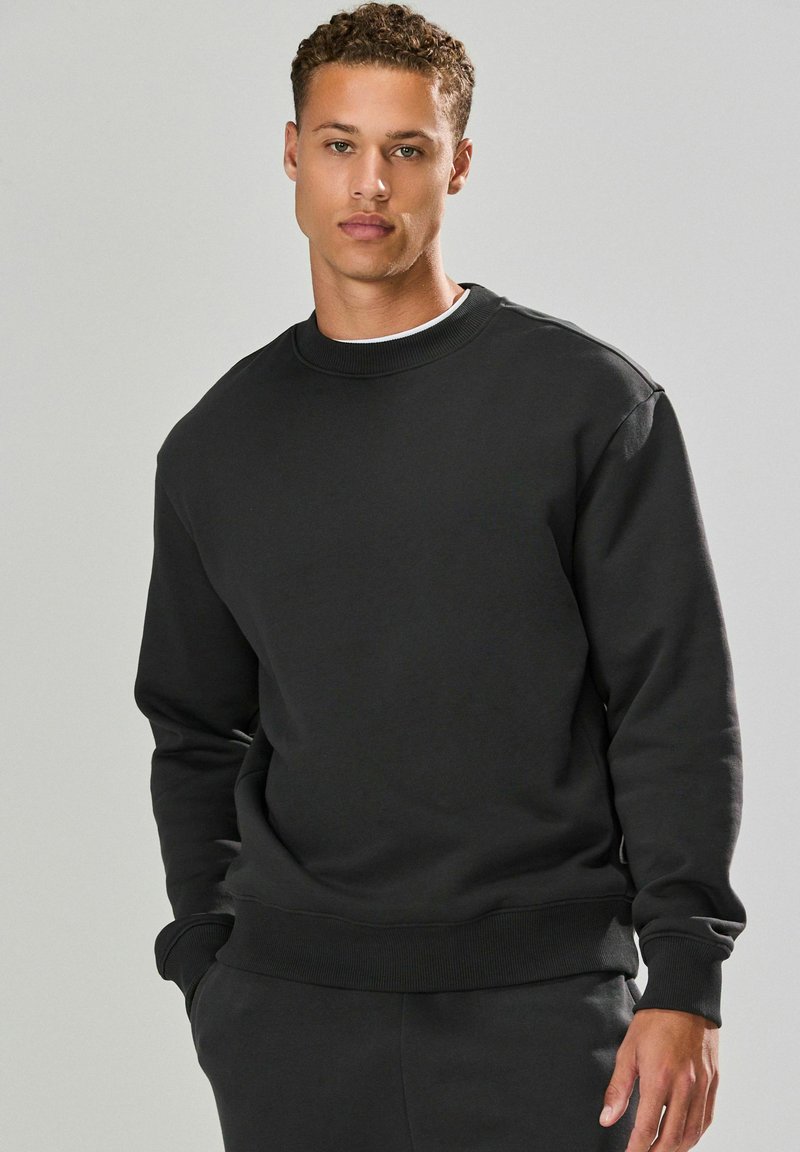 Junger Mann mit kurzen lockigen Haaren, der einen schlichten schwarzen Pullover und schwarze Hosen trägt, steht mit einer Hand in der Tasche vor einem grauen Hintergrund.