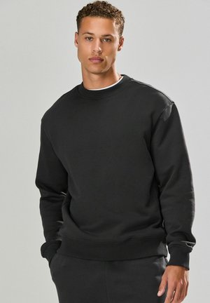 Ung mand med krøllet hår iført en sort sweatshirt og matchende bukser, der står mod en lys grå baggrund med den ene hånd i lommen.