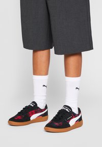 Sneakers Puma in suede nero, tessuto a quadri rossi, dettagli bianchi e suola in gomma marrone. Indossati con calze bianche con logo nero.