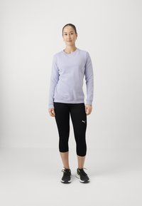 Lila Sweatshirt mit Rundhalsausschnitt, schwarze, cropped Leggings und schwarze Sportschuhe mit neonfarbigen Akzenten, vor einem weißen Hintergrund stehend.