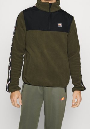 Mężczyzna ubrany w oliwkowo-zielony polar z czarnym panelem na klatce piersiowej oraz z logo marki, w połączeniu z dopasowanymi oliwkowo-zielonymi spodniami dresowymi.