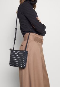 Svart crossbody-väska med prikkigt mönster och rektangulär form, med en slät yta och en lång, justerbar rem.