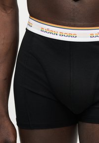 Μαύρα μποξεράκια με λευκή μέση που έχει ρίγες του ουράνιου τόξου και το κείμενο "BJÖRN BORG", φορεμένα από ένα άτομο με σκούρο δέρμα.