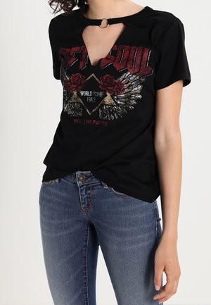 Femme portant un t-shirt graphique noir avec motif d'aile et de rose, encolure clé avec détail anneau, et jean bleu sur fond uni.