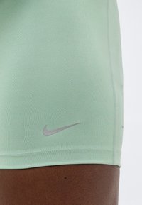 Κοντινή λήψη μηρού που φοράει ανοιχτό πράσινο σορτς Nike με ανακλαστικό λογότυπο σε λείο ύφασμα, με απλό φόντο.