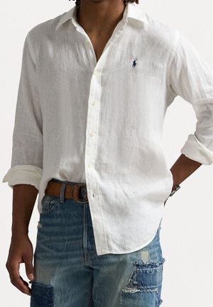 Man draagt een wit linnen overhemd met knopen en opgerolde mouwen en blauwe patchwork jeans met een bruine riem.