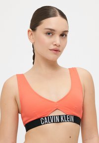 Korallfarbene Ripp-Bralette mit V-Ausschnittdesign, featuring einen schwarzen Calvin Klein Logo-Unterbund und einem Cut-Out-Detail in der Mitte.