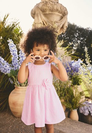 Joven niña con un vestido rosa a rayas ajusta unas gafas de sol con forma de flor, de pie al aire libre junto a grandes jarrones con plantas moradas y verdes.