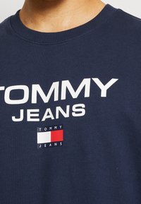 Námořnická modrá bavlněná mikina se silným bílým textem "TOMMY JEANS" a logem s červenými, bílými a modrými akcenty. Hladký ribovaný výstřih.