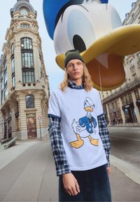 Bijela majica s plavim detaljima koja prikazuje grafiku Donalda Daka, složena preko košulje na kockice s dugim rukavima; na otvorenom, uz veliki balon.