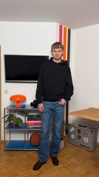 Νεαρός άνδρας με μαύρη φούτερ με κουκούλα και μπλε τζιν στέκεται σε δωμάτιο με τηλεόραση τοίχου, πορτοκαλί λάμπα, μπάλα του μπάσκετ και μεταλλική βαλίτσα με την ετικέτα Tropicana.