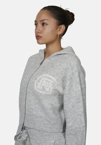 Szara bluza z kapturem na zamek, uszyta z miękkiego materiału, z prążkowanym pasem u dołu, naszywką z logo oraz detalem w postaci sznurka. Projekt obejmuje kaptur i zamek z przodu.