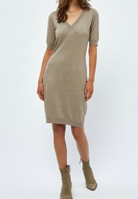 Robe en maille beige avec un col en V, manches courtes et une silhouette ajustée, arrivant aux genoux. Portée avec des bottines taupe arrivant à la cheville.