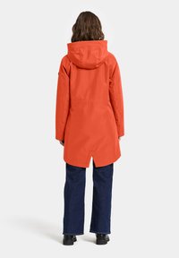 Veste imperméable orange avec capuche ; présente un ourlet arrière plus long et des coutures horizontales près de la taille, associée à un jean bleu foncé et des bottes noires.
