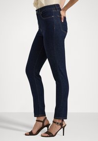 Slim-fit dark blue jeans worn by a person standing, paired with brown high-heeled sandals and a light-colored top.