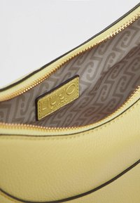 Intérieur d'un sac en cuir jaune avec une fermeture éclair. La doublure marron présente un motif ondulé subtil. Étiqueté avec une plaque rectangulaire dorée.