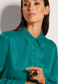 Vrouw in een teal satin button-up shirt, grote zwarte hoops oorbellen en een zwarte bloemenring aan haar rechterhand.