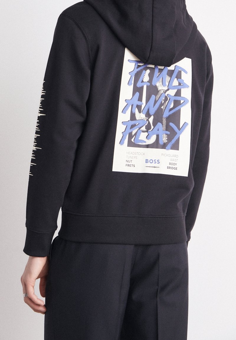 Il maglione con cappuccio nero presenta una stampa grafica sulla schiena con lettering blu audace, polsini a coste e una vestibilità rilassata.
