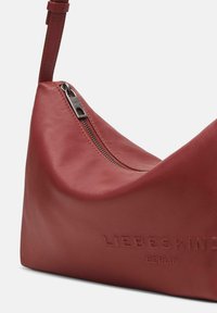 Borsa a tracolla in pelle rossa con forma curva, presenta una chiusura con zip e dettaglio del logo impresso. Texture liscia e tracolla regolabile.