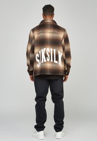 Brun, rutiga flanelljacka med vit "SIKSILK" logotyp på ryggen; svarta jeans; vita sneakers; texturen verkar mjuk och varm.