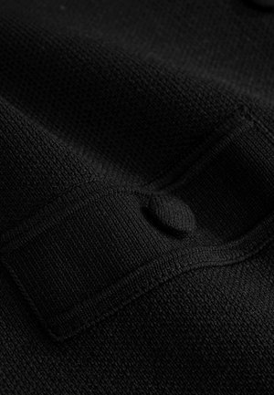 Melnas teksturētas auduma ar pinuma rakstu, ar pogu un atloka detaļu, rādot gludu apdari un izturīgu konstrukciju.