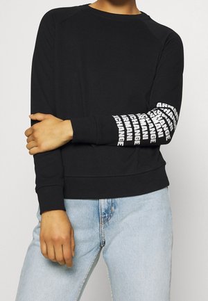 Osoba w czarnej długiej koszulce z powtarzającym się białym napisem „Armani Exchange” na prawym rękawie oraz jasnoniebieskich dżinsach.