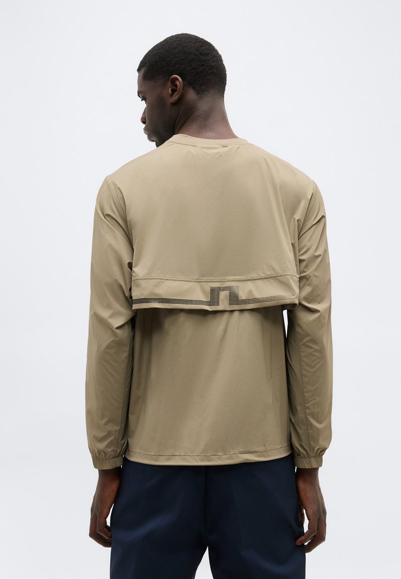 Muž s béžovou bundou s dlhými rukávmi a detailom prekrytia na zadnom rozparku, v tmavých nohaviciach, stojaci pred jasným svetlým pozadím.