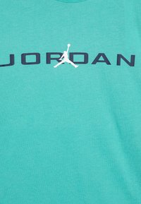 Turquoise katoenen T-shirt met een ronde hals; bevat "JORDAN" in vetgedrukte zwarte letters en een witte Jumpman-logo, gecentreerd boven de tekst.