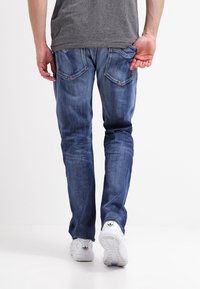 Tajta mörkblå jeans med en slät textur, subtila blekningar och dubbla bakfickor. Kombinerat med vita sportskor.