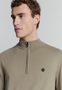 Olijfgroene trui met een hoge kraag en ritssluiting. Beschikt over een gestructureerde breiwerk en een hexagonaal logo patch op de borst.