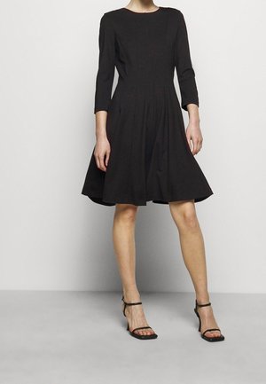 Woman wearing a knee-length black dress with three-quarter sleeves and black strappy heeled sandals, standing against a plain background.