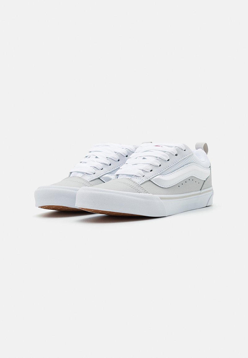 Zapatillas blancas con acentos grises, de lona texturizada y materiales de caucho, con punta redonda y suelas planas, que presentan un diseño clásico de cordones.