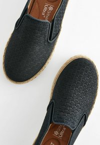 Schwarze gewobene Schlupfschuhe mit flacher Espadrille-Sohle aus Jute und braunem gepolstertem Fußbett. Sie verfügen über ein glattes, strukturiertes Obermaterial.