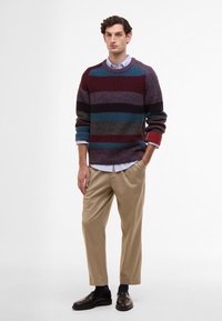 Multicolored knitted sweater with ribbed texture, featuring horizontal stripes in burgundy, blue, purple, and gray. Paired with beige trousers.